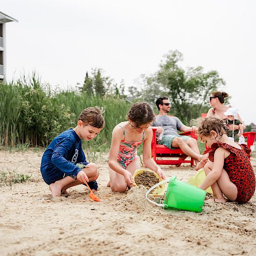 Three children play in the sand with buckets and shovels while two adults sit on red chairs by the beach.