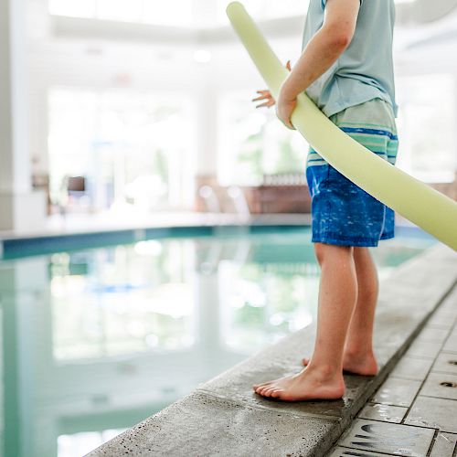 A child stands next to an indoor pool, holding a yellow pool noodle, wearing blue swim trunks and looking at the water.