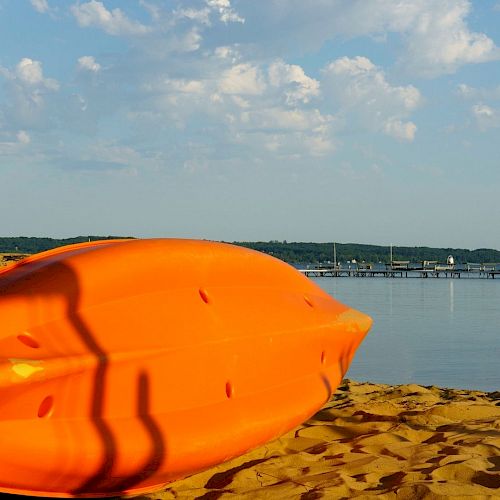 An orange kayak lies on the sandy beach with a calm lake, a dock, and a boat in the background on a sunny day, with scattered clouds in the sky.