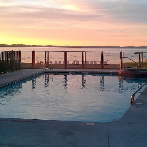 A swimming pool and hot tub are surrounded by a fence, with a view of the beach and ocean at sunset.