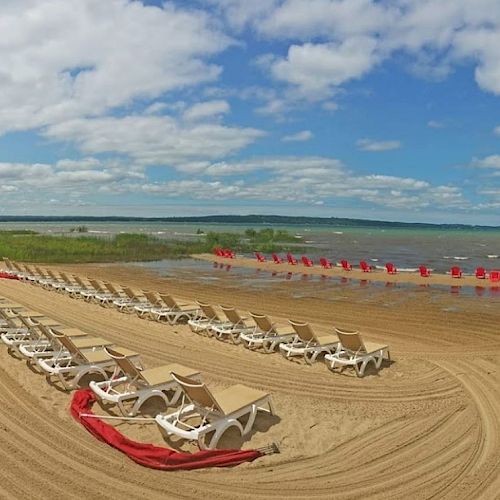 A sandy beach with rows of white lounge chairs, some red mats, and a view of a lake or sea under a blue sky with scattered clouds.