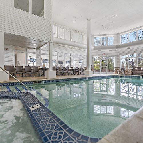 This image shows an indoor swimming pool area with large windows, natural light, seating, and a hot tub section.