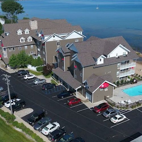 An aerial view of a lakeside resort with parking, a swimming pool, beach area with red umbrellas, and multiple buildings near a road.