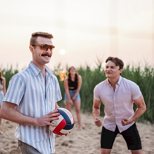 People are playing beach volleyball with excitement and enjoyment, surrounded by tall grass and a serene sunset in the background.