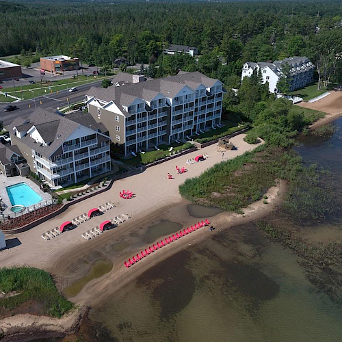 Aerial view of a beachside resort with buildings, parking lot, outdoor pool, and beachfront area with chairs and umbrellas, next to calm water.