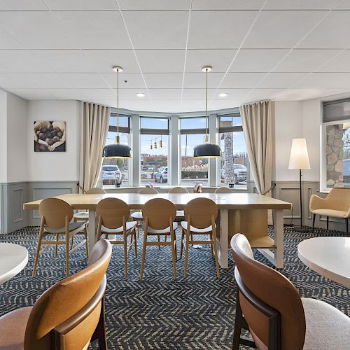 The image shows a modern, well-lit dining area with a long table and several chairs, pendant lights, and large windows with curtains.