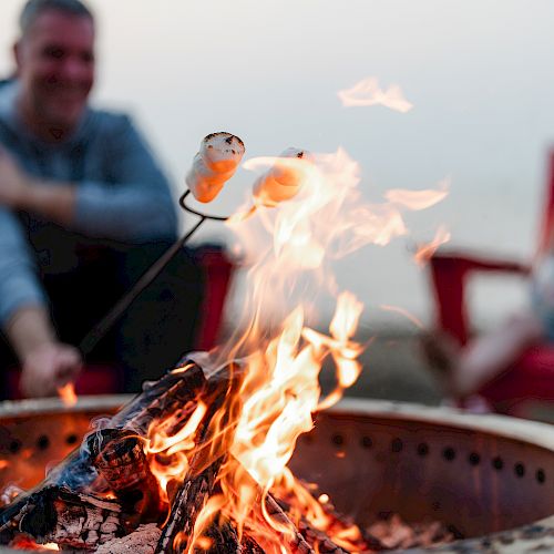 A person toasts marshmallows over a fire pit while another sits in the background, with red chairs around the fire.