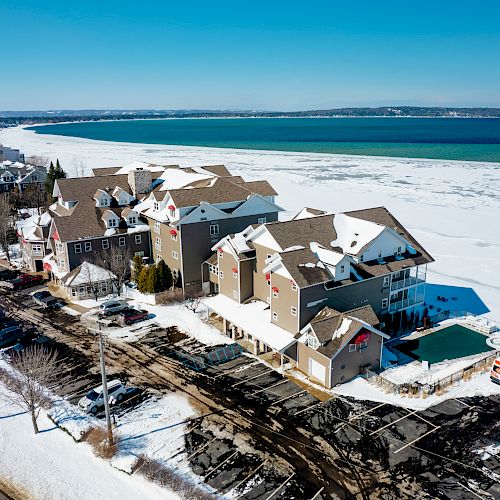 A snowy coastal town with buildings near a partially frozen body of water, clear skies, and a parking area with some cars.