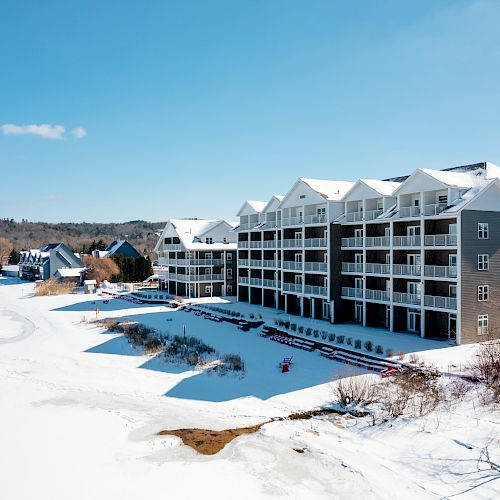 A large, snow-covered building with balconies, overlooking a frozen lake surrounded by other buildings and trees, under a clear blue sky.