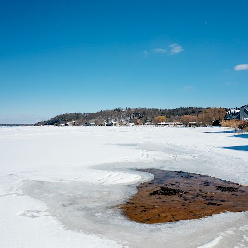 A snow-covered landscape with a partly frozen body of water, houses in the background, and under a clear blue sky, ending the sentence.