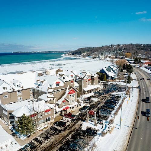 Aerial view of a snowy coastal town with colorful buildings, a road with cars, and a nearby partially frozen body of water under a clear blue sky.