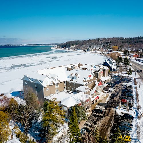 Aerial view of a snowy coastal town with buildings, a road, and a frozen body of water. Trees and vehicles are visible. The sky is clear.