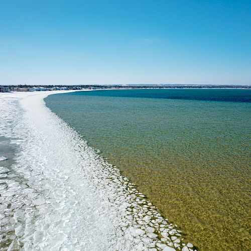 The image shows a coastline with a striking contrast between frozen, icy sections and open, clear water under a bright blue sky.