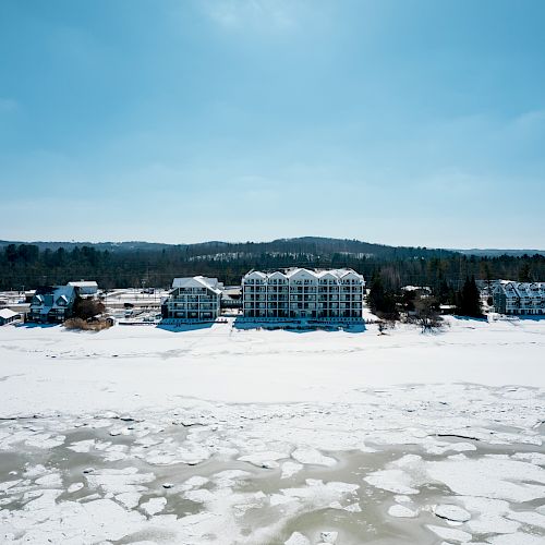 A line of buildings sits by a frozen lake, surrounded by snow and overlooked by forested hills under a clear blue sky.