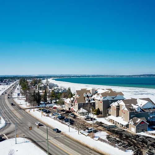 A snowy coastal town with a main road, buildings, and cars along a frozen body of water under a clear blue sky are visible in the image.