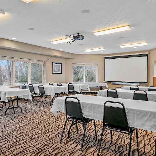 A conference room with multiple tables and chairs, a projector, and a screen, set up for a meeting or presentation, with windows on one side.