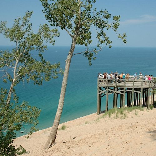 A scenic overlook with several people on a wooden platform viewing a vast body of water, sandy terrain, and trees under a clear blue sky.