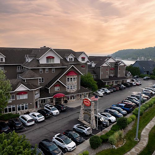 A large building, possibly a hotel or lodge, with many cars parked in front, next to a road during sunset with a scenic background.