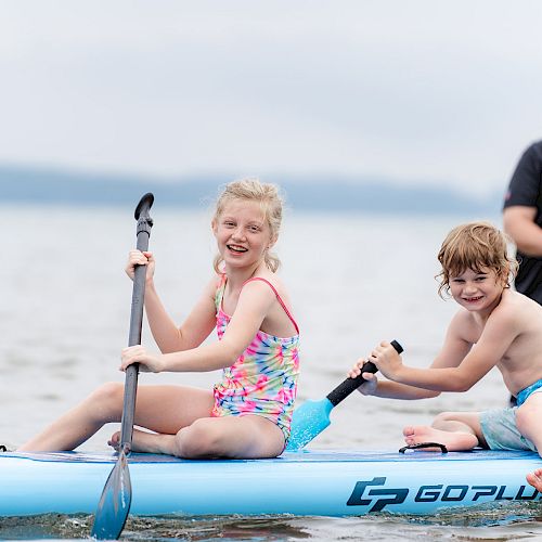 Two children paddle on a blue inflatable kayak, while an adult stands behind them in the water, smiling. They appear to be enjoying their time.