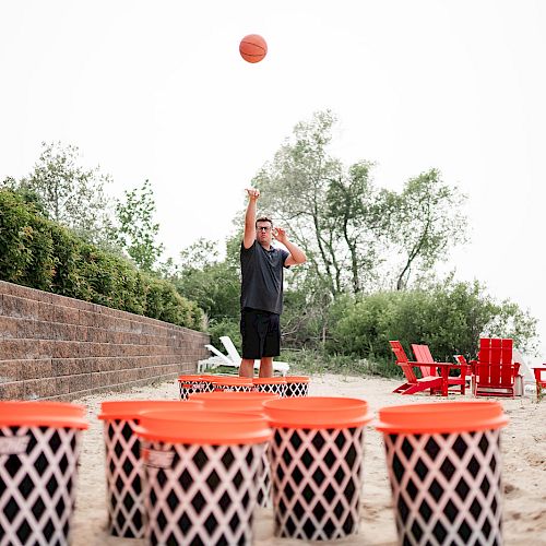 A person is playing a game with large buckets and an orange ball on a sandy area near greenery and red chairs.