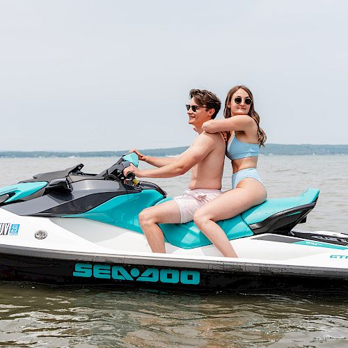 Two individuals are riding a Sea-Doo jet ski in a calm body of water; they are both wearing swimwear and sunglasses.