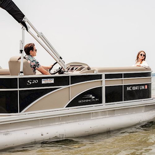 A person is steering a pontoon boat while another stands at the front, enjoying the view on calm waters.