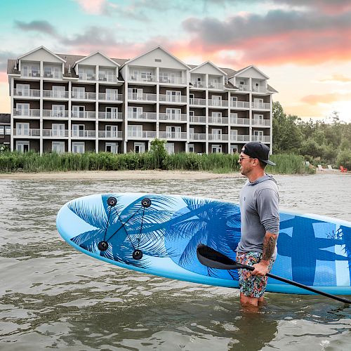 A man stands in shallow water holding a blue paddleboard, with a multi-story lakeside building and a colorful sunset in the background.