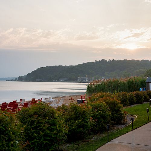 The image shows a serene beach setting with numerous lounge chairs, a calm ocean, lush greenery, and a winding path at sunset.