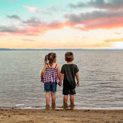 Two children holding hands stand at the edge of a beach, looking out at the ocean under a colorful sunset sky.