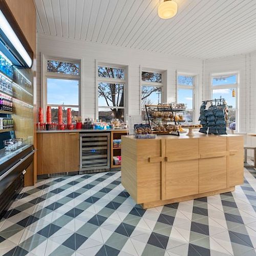 The image shows a modern, bright store interior with wooden shelves, a central island display, and large windows. Items are displayed on racks and shelves.