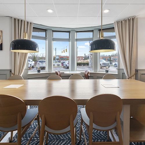 This image shows a modern, well-lit conference room with a long wooden table, chairs, two hanging lamps, large windows, and curtains.