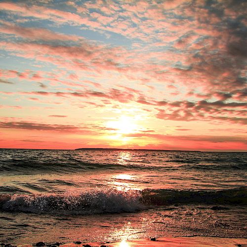 A beautiful sunset over the ocean with waves gently hitting the shore and scattered clouds in the sky.