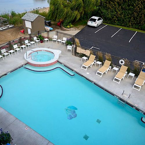 This image shows an outdoor swimming pool with a hot tub, lounge chairs, and a small parking lot next to it, located near a body of water.