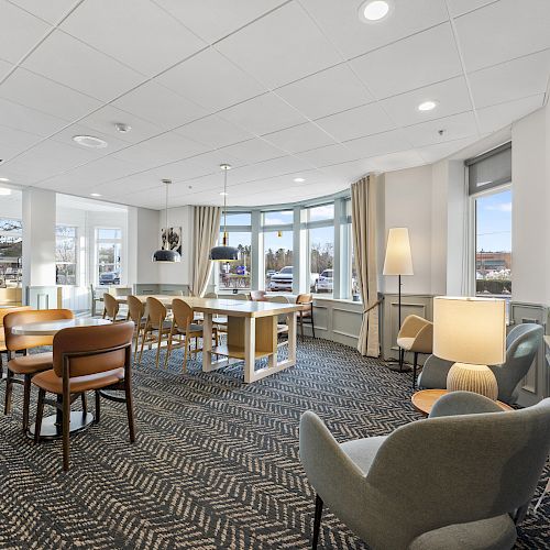 A modern, well-lit dining area with wooden tables and chairs, large windows, pendant lighting, and a patterned carpet.