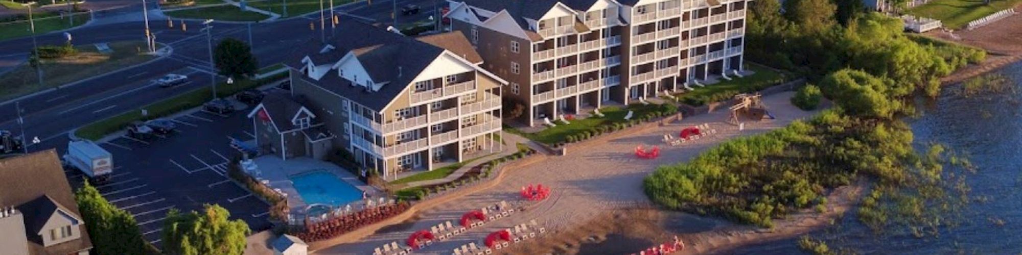 A beachfront hotel with a pool, beach chairs, and umbrellas. Adjacent road and buildings in the background, surrounded by greenery, ending the sentence.