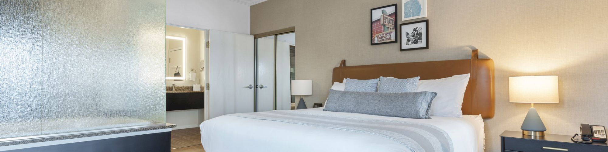 A modern bedroom features a large bed, nightstands with lamps, and a glass-paneled bathroom area visible from the room.