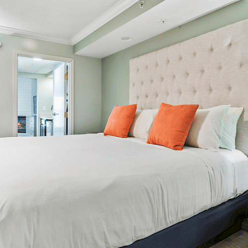 A cozy bedroom with a large bed, orange pillows, side tables, a lamp, and an open door leading to another room.