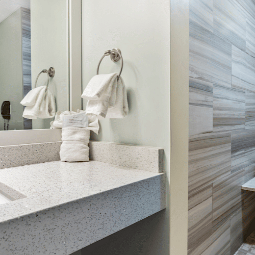 A modern bathroom with a countertop, sink, mirrors, towels on a hook, and a walk-in shower area with a tiled wall and a small bench.