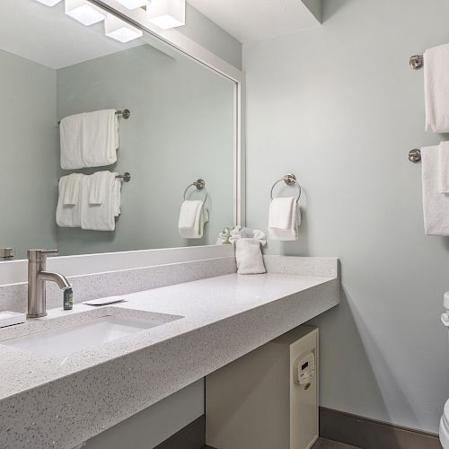 This image shows a modern bathroom with a sink, mirror, towels on racks, a toilet, and a shower area. The design is minimalistic and clean.