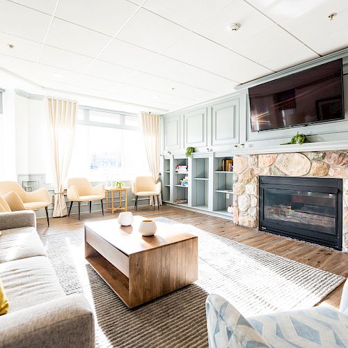 A cozy living room with sofas, armchairs, a wooden coffee table, large fireplace, built-in bookshelf, and a TV mounted above the mantle.