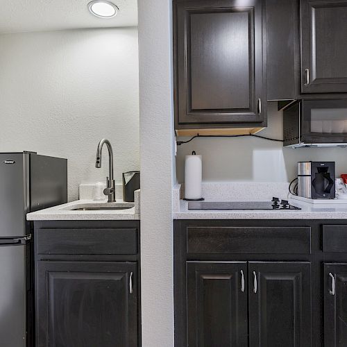 A compact kitchen with dark cabinets, a small fridge, sink, microwave, stovetop, coffee maker, and various items on the countertop.