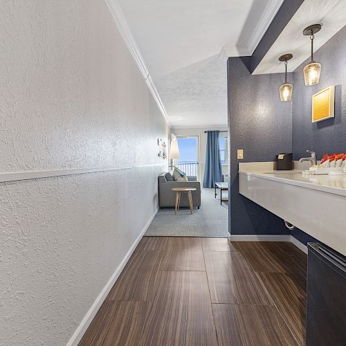 A modern hotel room entrance with a wood-floored hallway, countertop and desk, leading to a bright living area with a window view.