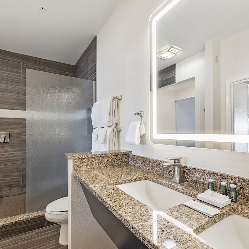 A modern bathroom with a granite countertop, double sinks, a large mirror with lights, a toilet, and a glass-enclosed shower.