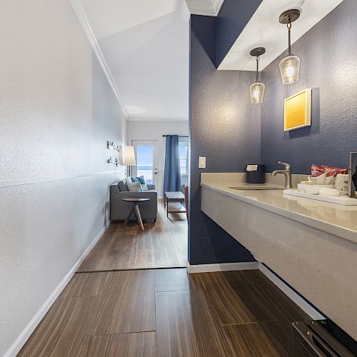 A modern hotel room features a kitchenette, pendant lighting, a sofa, and a window with curtains, leading to a sitting area in the distance.