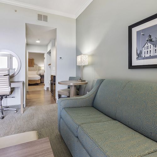 A modern hotel room features a sofa, desk with a chair, round table with two chairs, lamp, and photo on wall. Bedroom visible in the background.