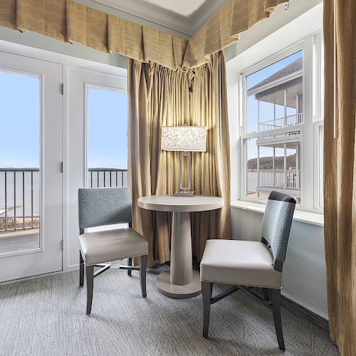 A cozy corner with a round table and two chairs near windows, featuring beige curtains and a lamp, offering a view of an outdoor balcony.