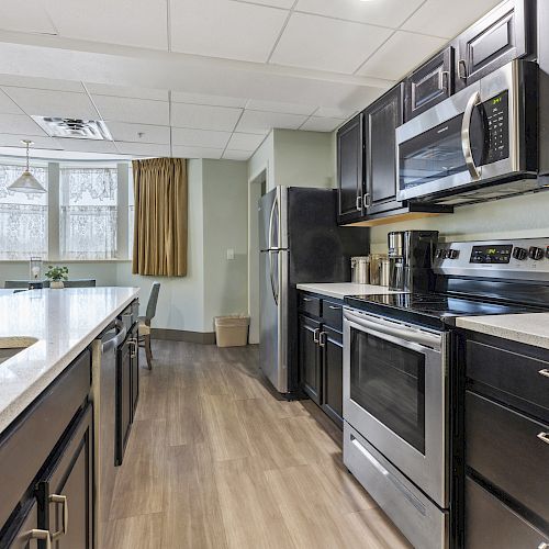 A modern kitchen with dark cabinets, stainless steel appliances, a center island, and a small dining area near a large window with curtains.