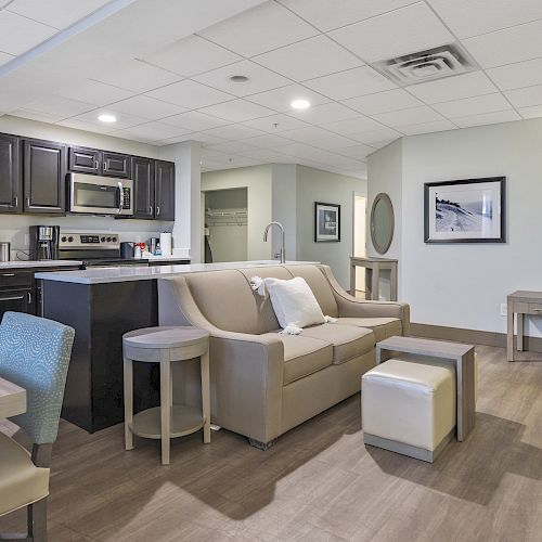 A modern living area with a couch, coffee table, armchair, and kitchen featuring stainless steel appliances and wooden cabinets is shown.