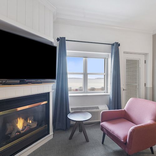 A cozy room features a fireplace, a TV, a pink armchair, a small table, and a window with blue curtains overlooking a scenic view.