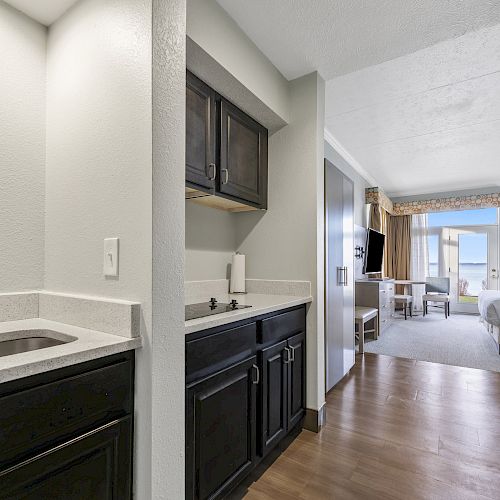 A hotel room with a small kitchenette, dark cabinets, sink, and white countertops leading into a bedroom with a bed and window view.